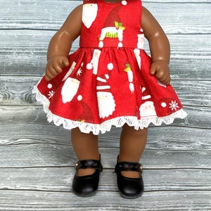 May include: A red sleeveless dress with a white lace trim and a pattern of white Santas and gnomes. The dress is worn by a doll with brown skin and black shoes.
