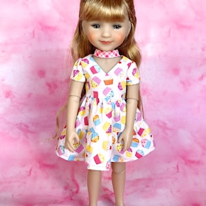 May include: A doll with blonde hair wearing a white dress with a cupcake pattern and a pink and white checkered collar. The doll is wearing white shoes and is standing on a pink background.