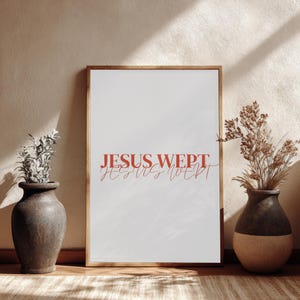 May include: A framed print with the words "JESUS WEPT" in red script. The print is displayed on a wooden surface, flanked by two decorative vases with dried plants. The frame is gold.