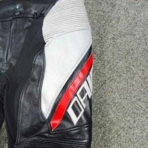 Może przedstawiać: Czarne sk&oacute;rzane spodnie motocyklowe z białymi i czerwonymi akcentami. Spodnie posiadają biały panel z czerwonym paskiem i napisem "DAINES" w kolorze białym. Obszar kolana ma żebrowane detale.
