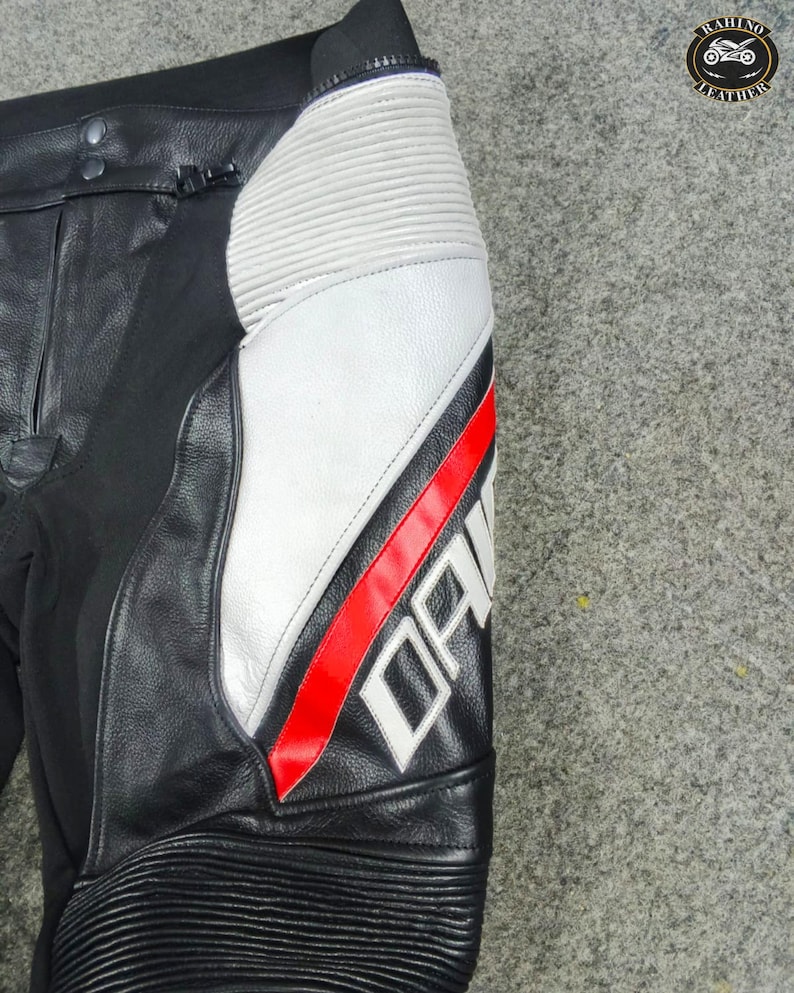 Może przedstawiać: Czarne sk&oacute;rzane spodnie motocyklowe z białymi i czerwonymi akcentami. Spodnie posiadają biały panel z czerwonym paskiem i napisem "DAINES" w kolorze białym. Obszar kolana ma żebrowane detale.