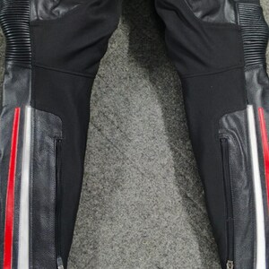 Może przedstawiać: Czarne spodnie motocyklowe ze sk&oacute;rzanymi panelami i czerwono-białymi paskami po bokach. Spodnie mają zamki błyskawiczne przy kostkach i prążkowane detale na kolanach.