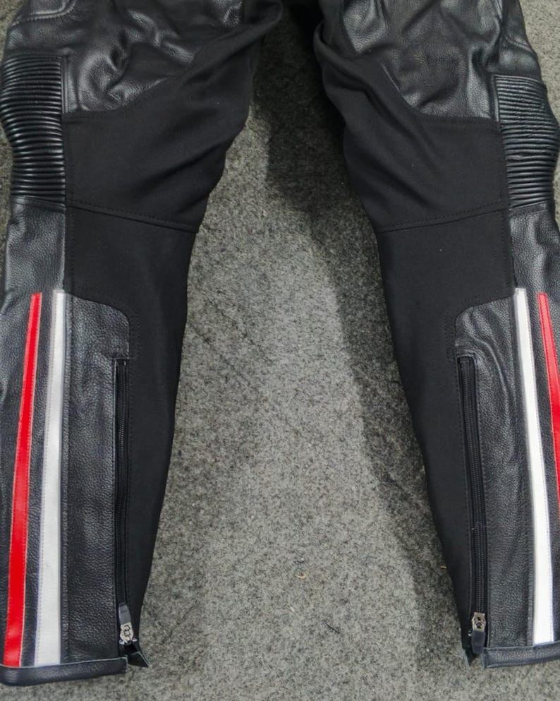 Może przedstawiać: Czarne spodnie motocyklowe ze sk&oacute;rzanymi panelami i czerwono-białymi paskami po bokach. Spodnie mają zamki błyskawiczne przy kostkach i prążkowane detale na kolanach.