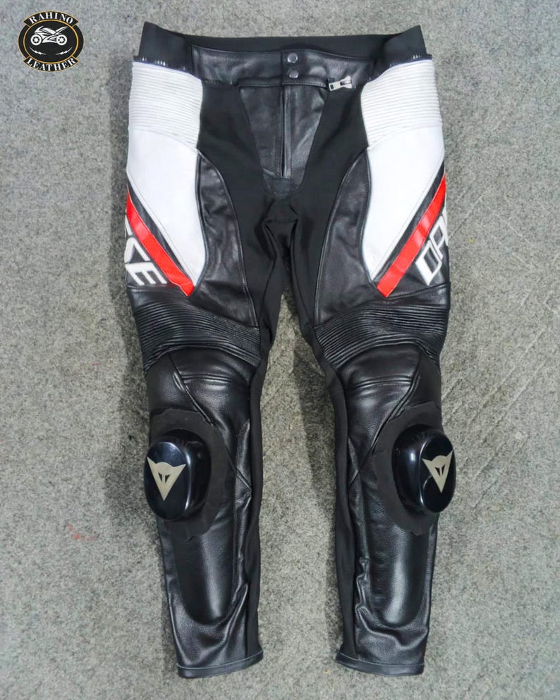 Może przedstawiać: Czarne i białe sk&oacute;rzane spodnie motocyklowe z czerwonymi akcentami. Spodnie posiadają slidery kolanowe i biały napis "DAINESE". Projekt obejmuje białe panele po bokach i prążkowane detale nad kolanami. Spodnie mają zapięcie na zamek błyskawiczny.