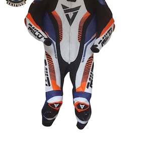 May include: A multi-coloured leather motorcycle racing suit in black, white, blue, and orange. The suit has a white central panel with a black triangle logo and the word "MILU" on the arms and legs. It features a full-length zip and is designed for protection.