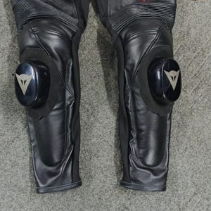 Może przedstawiać: Czarne sk&oacute;rzane spodnie motocyklowe z białymi i czerwonymi akcentami. Spodnie posiadają ochraniacze kolan z czarno-srebrnym logo. Sk&oacute;ra jest teksturowana, a spodnie mają prążkowane panele. Zaprojektowane do jazdy motocyklem.