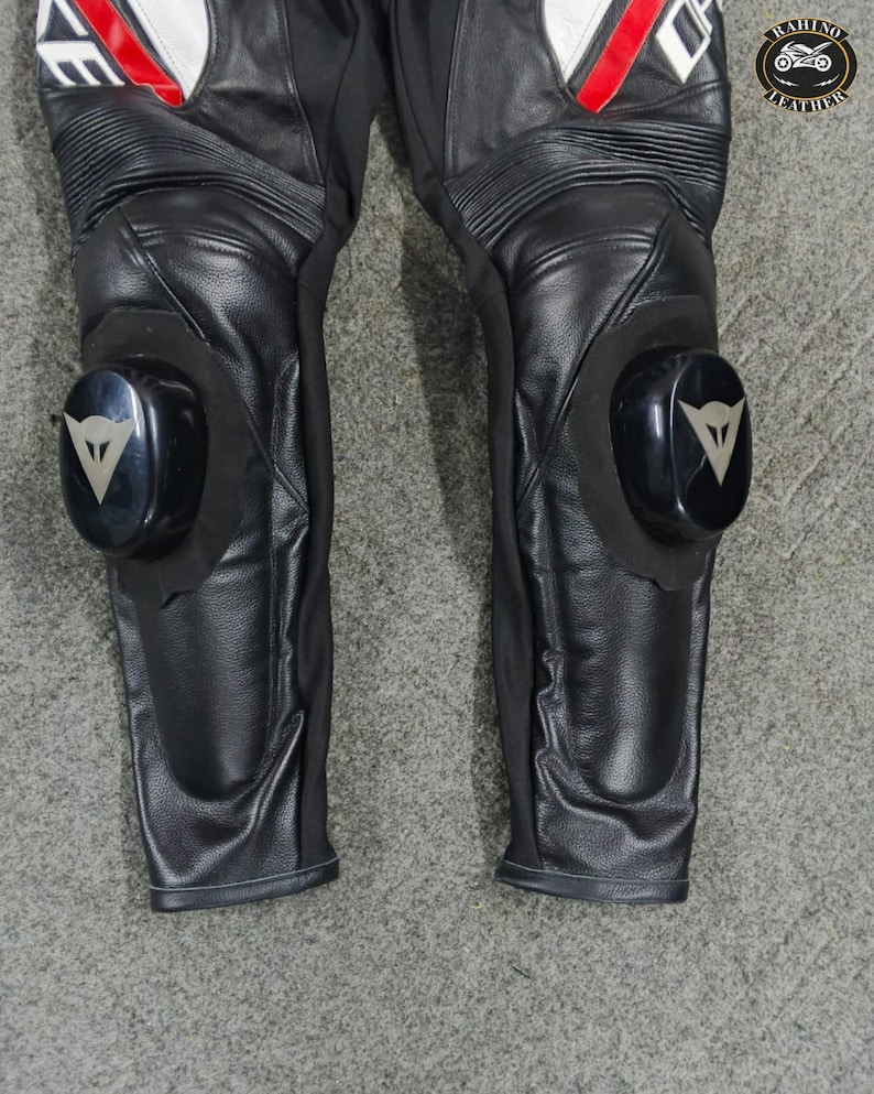 Może przedstawiać: Czarne sk&oacute;rzane spodnie motocyklowe z białymi i czerwonymi akcentami. Spodnie posiadają ochraniacze kolan z czarno-srebrnym logo. Sk&oacute;ra jest teksturowana, a spodnie mają prążkowane panele. Zaprojektowane do jazdy motocyklem.