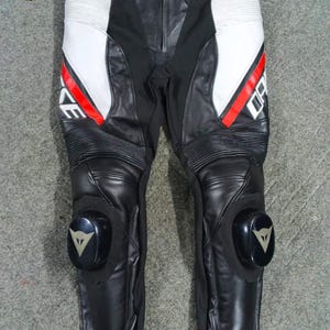 Może przedstawiać: Czarne i białe sk&oacute;rzane spodnie motocyklowe z czerwonymi akcentami. Spodnie posiadają ochraniacze kolan i nazwę marki "DAINESE" w kolorze białym. Projekt obejmuje białe panele i żebrowane sekcje. Spodnie przeznaczone do jazdy motocyklem.