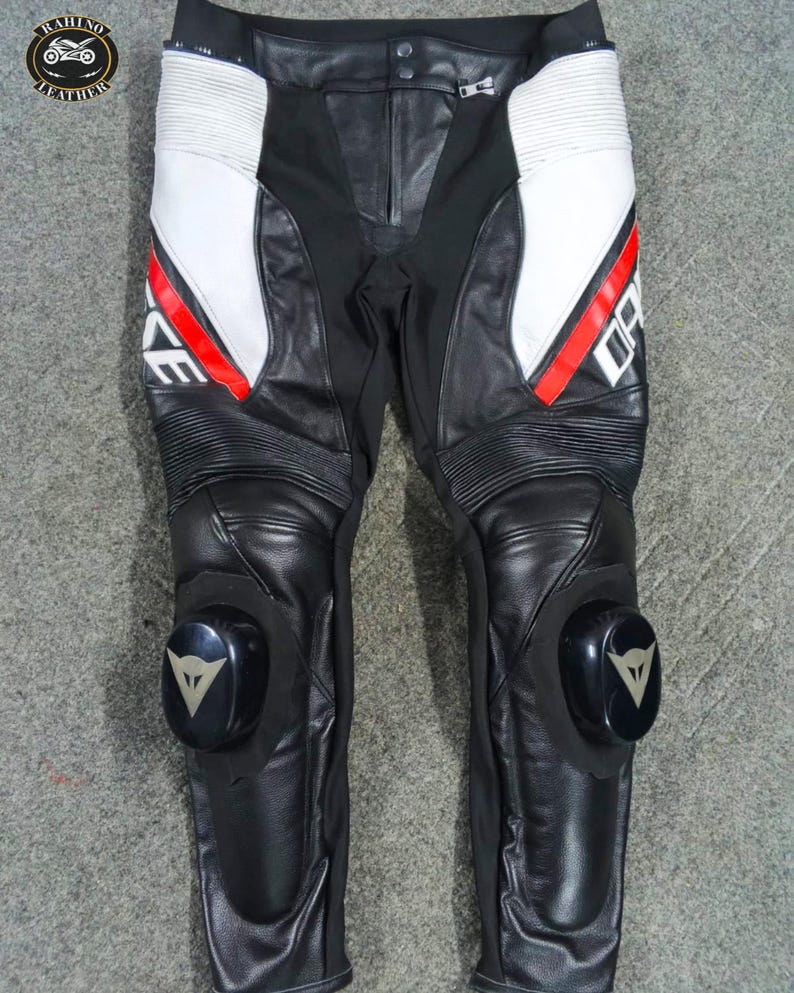 Może przedstawiać: Czarne i białe sk&oacute;rzane spodnie motocyklowe z czerwonymi akcentami. Spodnie posiadają ochraniacze kolan i nazwę marki "DAINESE" w kolorze białym. Projekt obejmuje białe panele i żebrowane sekcje. Spodnie przeznaczone do jazdy motocyklem.
