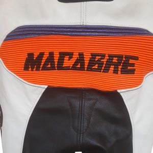 May include: Close-up of motorcycle racing trousers. The trousers are white, black, and orange with the word "MACABRE" printed in black on the orange section. The trousers have a leather appearance.