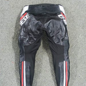 Może przedstawiać: Czarne sk&oacute;rzane spodnie motocyklowe z białymi i czerwonymi akcentami. Spodnie mają napis "Dainese" w kolorze białym. Spodnie mają zamki błyskawiczne na dolnych nogawkach oraz białe, czerwone i białe paski po bokach.