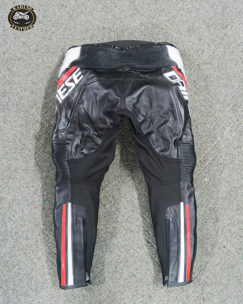 Może przedstawiać: Czarne sk&oacute;rzane spodnie motocyklowe z białymi i czerwonymi akcentami. Spodnie mają napis "Dainese" w kolorze białym. Spodnie mają zamki błyskawiczne na dolnych nogawkach oraz białe, czerwone i białe paski po bokach.