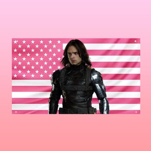 May include: A pink and white American flag with a person in a black tactical vest in front of it. The person has long brown hair and is looking towards the viewer. The background is a light pink colour.