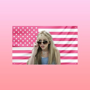 May include: A pink American flag with white stars and stripes is in the background. A person with long blonde hair is wearing sunglasses and a blue top. The person is looking at the camera.