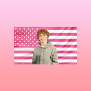 May include: A person wearing a gray hoodie stands in front of a pink American flag. The flag has pink stripes and white stars. The background is a gradient of pink.