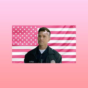 May include: A man in a police uniform with a pink bow on his head stands in front of a pink American flag. The flag has pink stripes and white stars. The man is wearing a black jacket with the name "Bradford" visible.