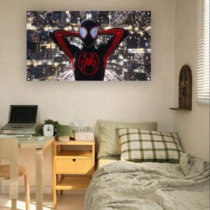 Puede incluir: Una impresión artística en lienzo enmarcada que presenta a un superhéroe con un traje negro y rojo con un emblema de araña. La obra de arte representa al personaje contra un telón de fondo urbano, con los brazos detrás de la cabeza.