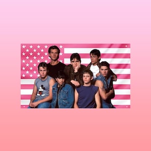 May include: A group of seven people posing in front of a pink American flag. The people are wearing various outfits, including denim jackets, tank tops, and t-shirts. The background is a solid pink color.