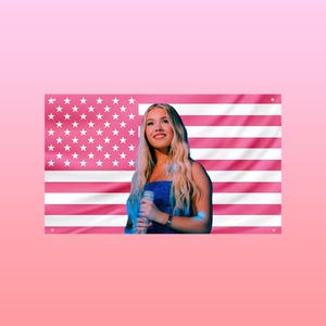 May include: A woman with long blonde hair wearing a blue sequined top, holding a microphone. Behind her is a pink American flag. The background is a soft pink.