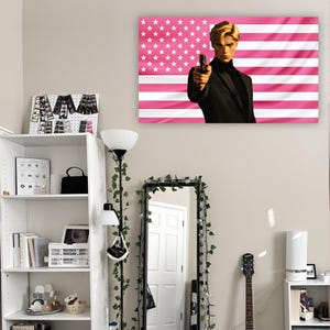 May include: A rectangular wall tapestry featuring a stylized illustration of a blonde person in a black suit holding a handgun, set against a background of horizontal pink and white stripes with a field of white stars in the upper left corner.