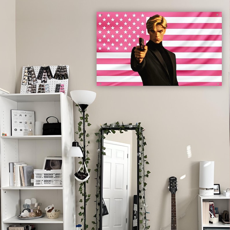 May include: A rectangular wall tapestry featuring a stylized illustration of a blonde person in a black suit holding a handgun, set against a background of horizontal pink and white stripes with a field of white stars in the upper left corner.