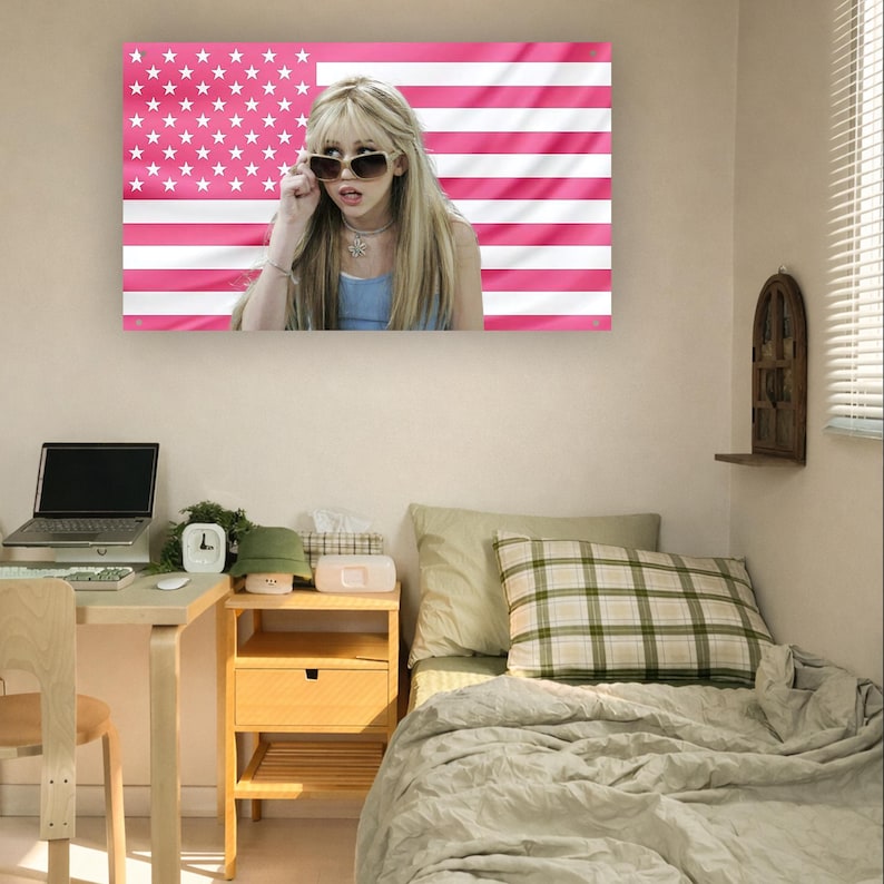 May include: A canvas print of a woman wearing sunglasses against a pink American flag. The room includes a desk with a laptop, a wooden bedside table, and a bed with a plaid cushion.