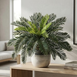 Könnte beinhalten: Eine künstliche Palme mit dunkel- und hellgrünen Wedeln in einem hellgrauen Keramiktopf. Der Topf steht auf einem hellen Holztisch, mit Designbüchern und einer geometrischen Metallskulptur. Dekoration für Innenräume.