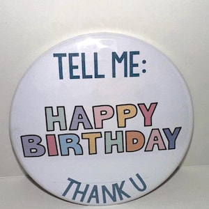 May include: A white, round button with the text "TELL ME: HAPPY BIRTHDAY THANK U" in various colors. The button has a glossy finish and a simple, bold font.