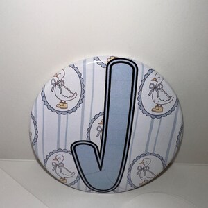 May include: A round button with a light blue letter "J" outlined in black. The background features a pattern of light blue stripes and duck illustrations within scalloped frames, creating a whimsical design.
