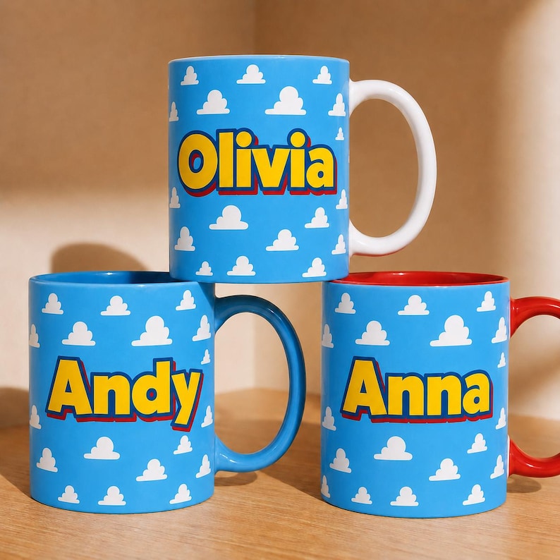 K&ouml;nnte beinhalten: Drei Keramiktassen mit hellblauem Hintergrund und einem sich wiederholenden wei&szlig;en Wolkenmuster. Jede Tasse zeigt einen personalisierten Namen in gro&szlig;en gelben Buchstaben mit rotem Umriss. Die Namen Olivia, Andy und Anna sind aufgedruckt.