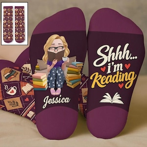 May include: A pair of purple socks featuring a cartoon illustration of a person with blonde hair reading a book while sitting among a stack of books. The text reads "Shhh... i'm Reading" and "Jessica". The socks have a dark purple heel and toe.