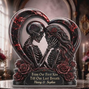 Può includere: Una scultura decorativa con due scheletri che si fronteggiano all'interno di una cornice a forma di cuore con motivi in marmo rosso e nero. La base è decorata con rose rosse e il testo: From Our First Kiss Till Our Last Breath Henry & Sophia.