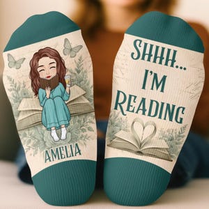 Könnte beinhalten: Ein Paar Socken in Blaugrün und Creme. Die linke Socke zeigt eine Illustration einer Person mit braunen Haaren, die ein Buch liest, Schmetterlinge und den Namen AMELIA. Die rechte Socke zeigt den Text SHHH... I'M READING über einem aufgeschlagenen Buch.