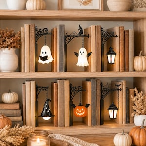 May include: A set of six decorative bookends featuring black metal wall-mount style brackets with hanging ornaments including white ghosts, a black witch hat, an orange jack-o'-lantern, and black lanterns, all displayed between rows of books.