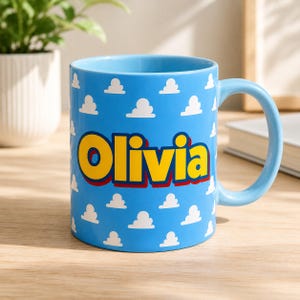 K&ouml;nnte beinhalten: Ein hellblauer Keramikbecher mit einem sich wiederholenden Muster aus kleinen wei&szlig;en Wolken. Der Name Olivia ist in gro&szlig;en, fetten gelben Buchstaben mit dunkelroter Umrandung in der Mitte des Bechers aufgedruckt, der einen passenden Henkel hat.