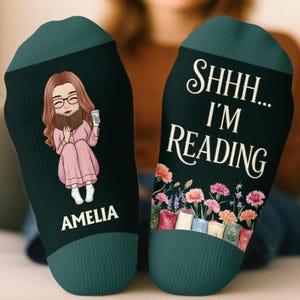 May include: A pair of dark green socks with ribbed cuffs. One sock features an illustration of a person with long brown hair reading a book while holding a cup, with the name AMELIA printed below. The other sock displays the text SHHH... I'M READING above a row of colorful books and flowers.