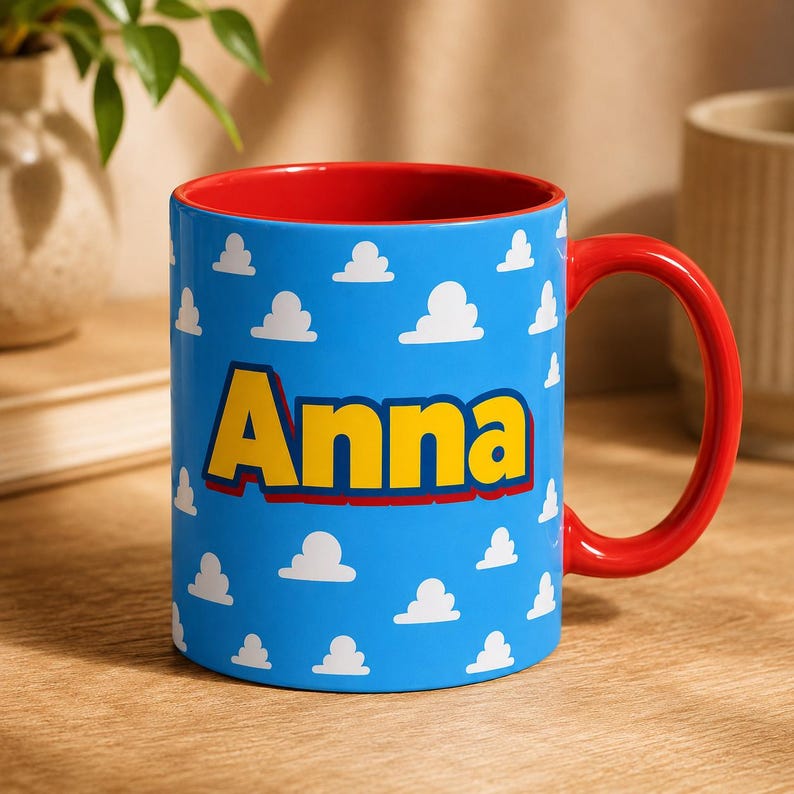 K&ouml;nnte beinhalten: Ein Keramikbecher mit einer leuchtend blauen Au&szlig;enseite, die mit einem sich wiederholenden Muster aus wei&szlig;en Wolken und dem Namen Anna in gro&szlig;en gelben Buchstaben mit dunkelroter Umrandung bedruckt ist. Der Becher hat ein rotes Inneres.
