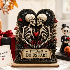Op de afbeelding: Een decoratieve plaquette met twee skeletten die tegenover elkaar staan met een rood hart ertussen. De namen Sophia en David staan bovenaan gegraveerd. Een banner aan de basis draagt de tekst Till Death DO US PART. Versierd met rode rozen.