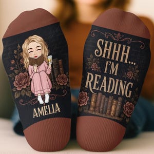 May include: A pair of dark socks with brown cuffs and heels. One sock features an illustration of a blonde person reading a book with the name AMELIA printed below. The other sock displays the text SHHH... I'M READING above a shelf of books.