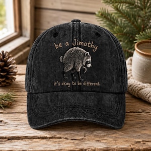 May include: A black washed-denim baseball cap featuring a graphic of a raccoon in the center. Above the raccoon, the text reads "be a Jimothy" and below the raccoon, the text reads "it's okay to be different" in a light brown, sans-serif font.