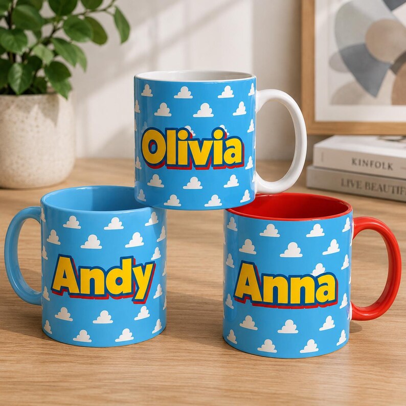 K&ouml;nnte beinhalten: Drei Keramiktassen mit hellblauem Hintergrund und einem sich wiederholenden wei&szlig;en Wolkenmuster. Jede Tasse zeigt einen personalisierten Namen in fetten gelben Buchstaben mit rotem Umriss: Olivia, Andy und Anna. Die Tassen haben wei&szlig;e oder rote Henkel.