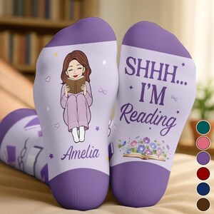 Könnte beinhalten: Hellviolette Socken mit einer Cartoon-Illustration einer lesenden Person, dem Namen Amelia und dem Text SHHH... I'M Reading über einem offenen Buch mit Blumen. Das Design enthält kleine Sterne und Schleifen auf einem lavendelfarbenen Hintergrund.