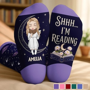 Könnte beinhalten: Ein Paar dunkelblaue Socken mit violetten Bündchen. Eine Socke zeigt eine blonde Person, die auf einer Mondsichel ein Buch liest, mit dem Namen AMELIA darunter. Die andere Socke zeigt den Text SHHH... I'M READING über einem aufgeschlagenen Buch.