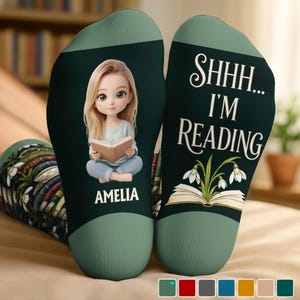 Könnte beinhalten: Ein Paar dunkelgrüne Socken mit einer Cartoon-Illustration eines blonden Mädchens, das ein Buch liest, mit dem Namen AMELIA darunter. Die andere Socke zeigt den Text SHHH... I'M READING über einem aufgeschlagenen Buch mit weißen Blumen.
