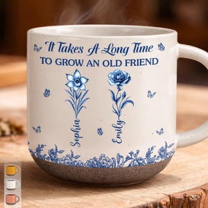 May include: A cream-colored ceramic mug with a speckled finish and a dark gray base. The mug features blue floral illustrations, butterflies, and the text "It Takes A Long Time TO GROW AN OLD FRIEND" with the names "Sophia" and "Emily" printed vertically.