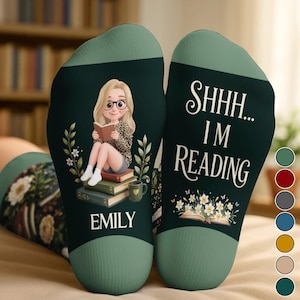 May include: A pair of dark green socks featuring a cartoon illustration of a blonde person with glasses reading a book while sitting on a stack of books, with the name EMILY printed below. The other sock displays the text SHHH... I'M READING.