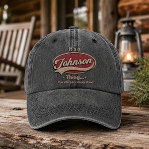 May include: A charcoal gray baseball cap with a distressed finish. The front features an embroidered red oval emblem with the text "It's a Johnson Thing... You Wouldn't Understand" in a cream-colored script and sans-serif font style.