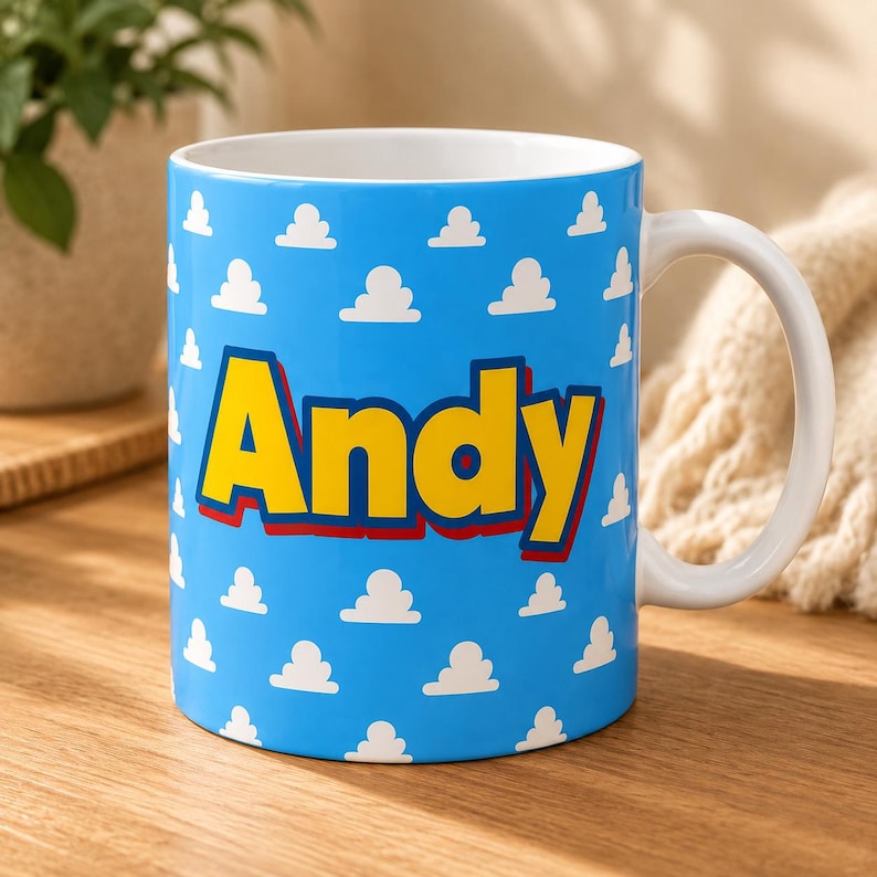 K&ouml;nnte beinhalten: Ein Keramikbecher mit hellblauem Hintergrund und einem sich wiederholenden Muster aus kleinen wei&szlig;en Wolken. Der Name Andy ist in gro&szlig;en, fetten gelben Buchstaben mit einer dicken roten Umrandung in der Mitte des Bechers aufgedruckt.