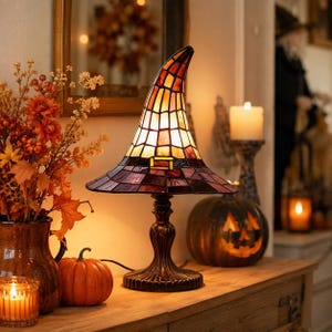 May include: A stained glass table lamp shaped like a pointed witch hat with a dark purple brim and an orange and yellow crown. The lamp features a dark metal base and a small decorative buckle detail on the front of the hat, emitting a warm glow.