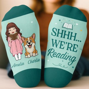 May include: A pair of light teal socks with dark teal cuffs. The left sock features an illustration of a person reading a book next to a corgi, with the names Amelia and Charlie. The right sock displays an open book icon and the text SHHH... WE'RE Reading.